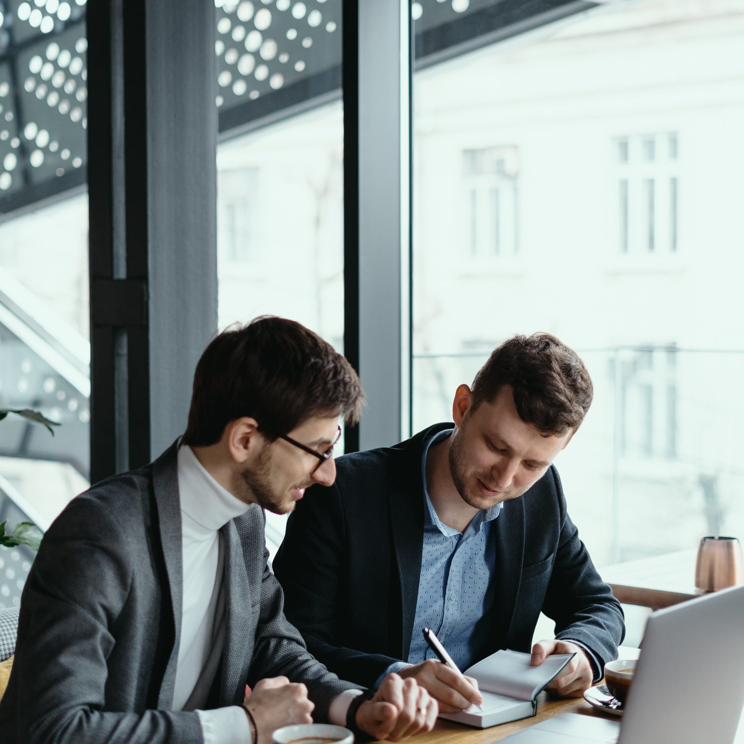 Two businessmen talking about new opportunities sitting with laptop at desk near large window, planning project, considering business offer, sharing ideas while drinking coffee together